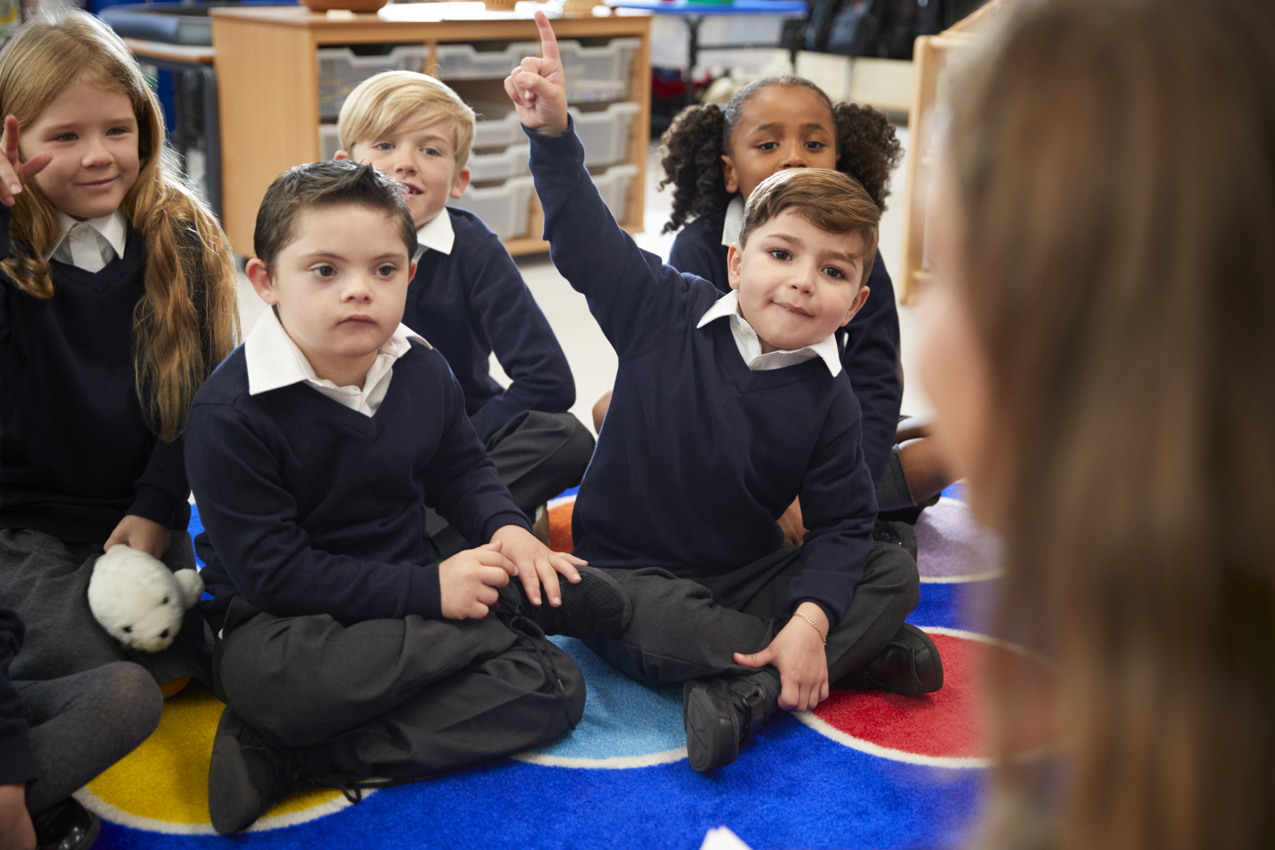 View over teacher's shoulder of school children sitting on the classroom floor raising hands to answer a question