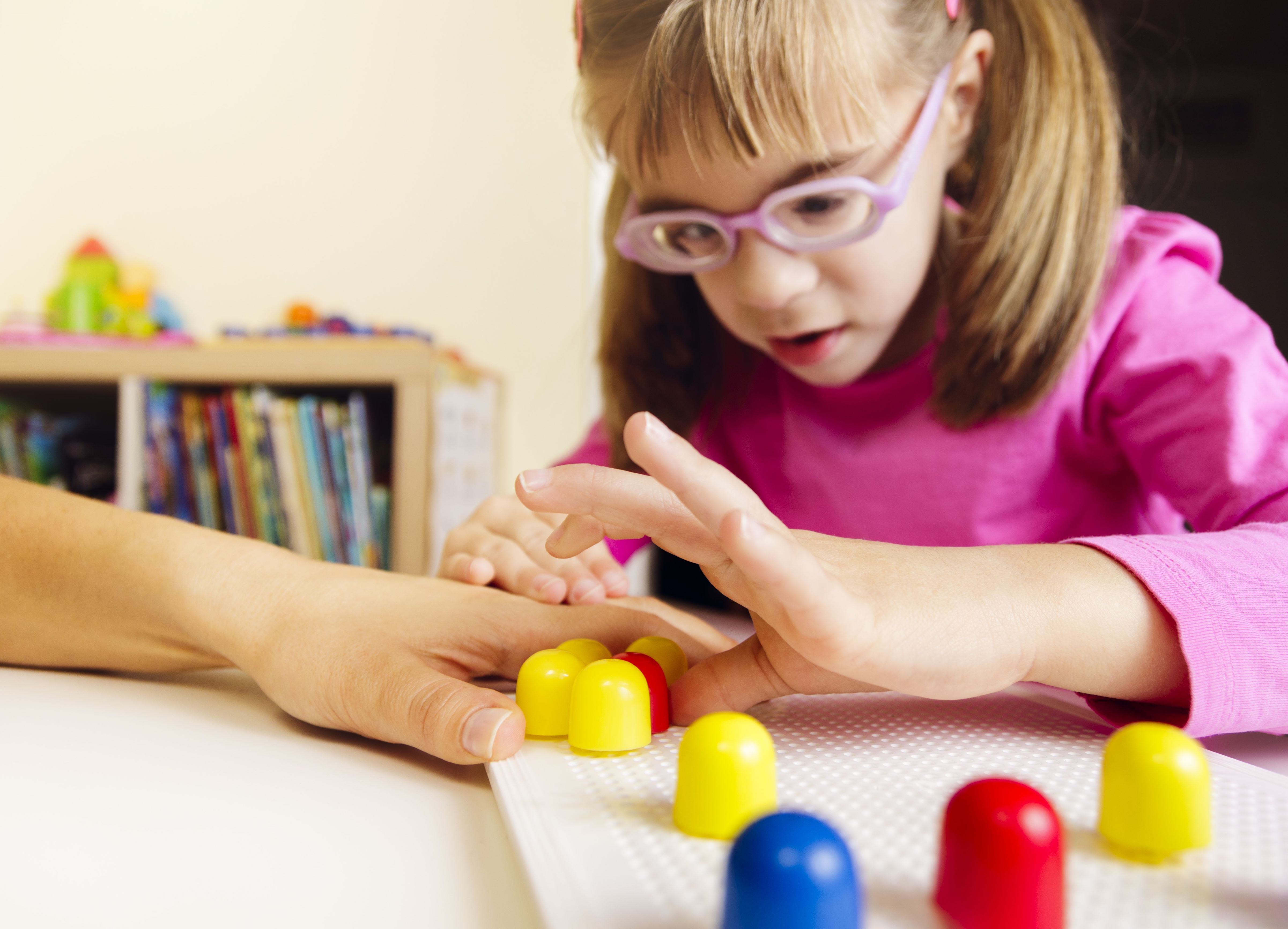 Young girl with vision impairment sticking mosaic pegs on a board with the help of her teacher
