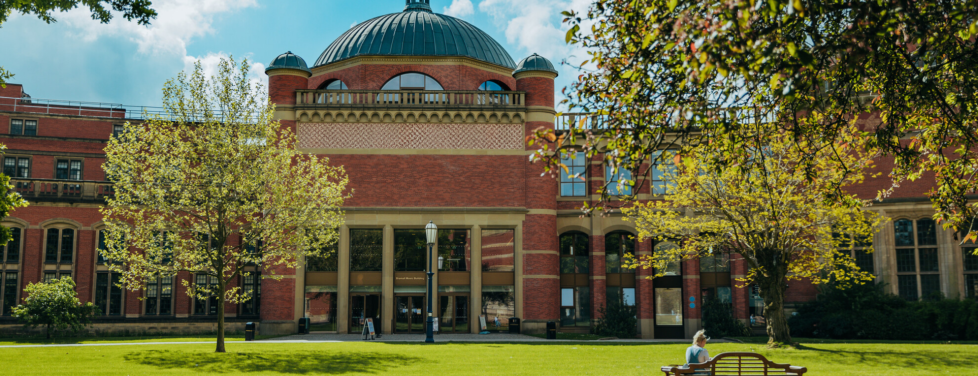 An image of Bramall Music Building, on a bright sunny day, with a student sitting by on a bench amongst some greenery.