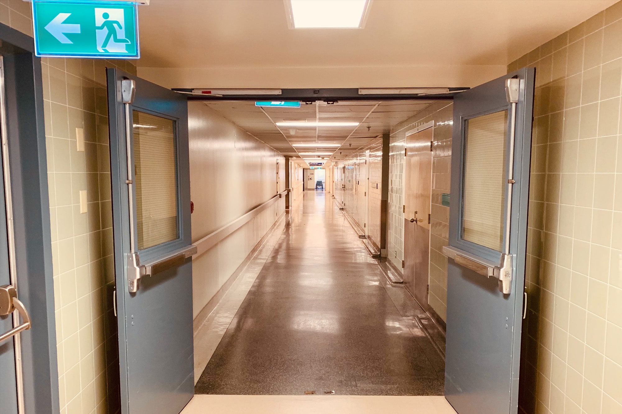 Long, empty hospital corridor with an exit sign pointing to a door on the left