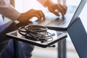 Male doctor hands typing on laptop computer keyboard next to a digital tablet and medical stethoscope on the desk.