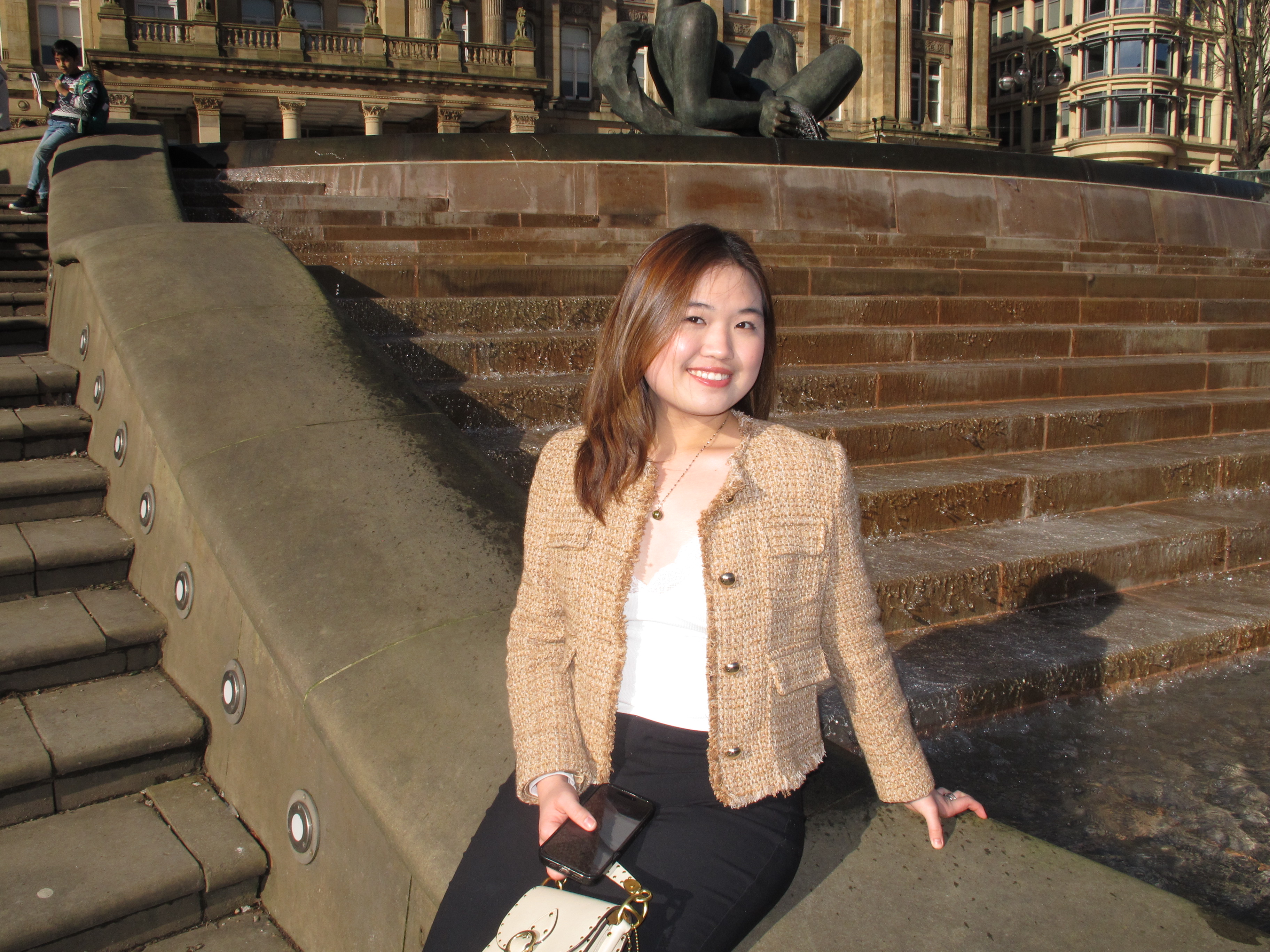 Jessica is sat outside Birmingham City Hall. She has long brown hair and is wearing a tan jacket, white top and black trousers. 