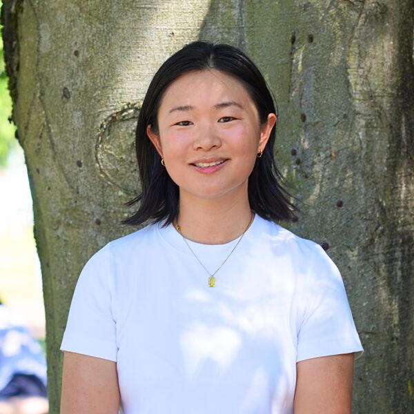 Joey wearing a white t-shirt and gold necklace stood in front of a tree smiling towards the camera.