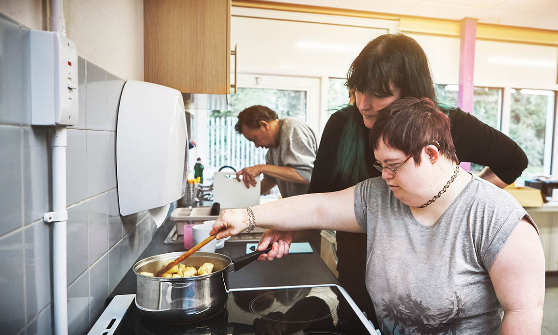 Young woman with Downes Syndrome stirs a pot on a stove, with her mother