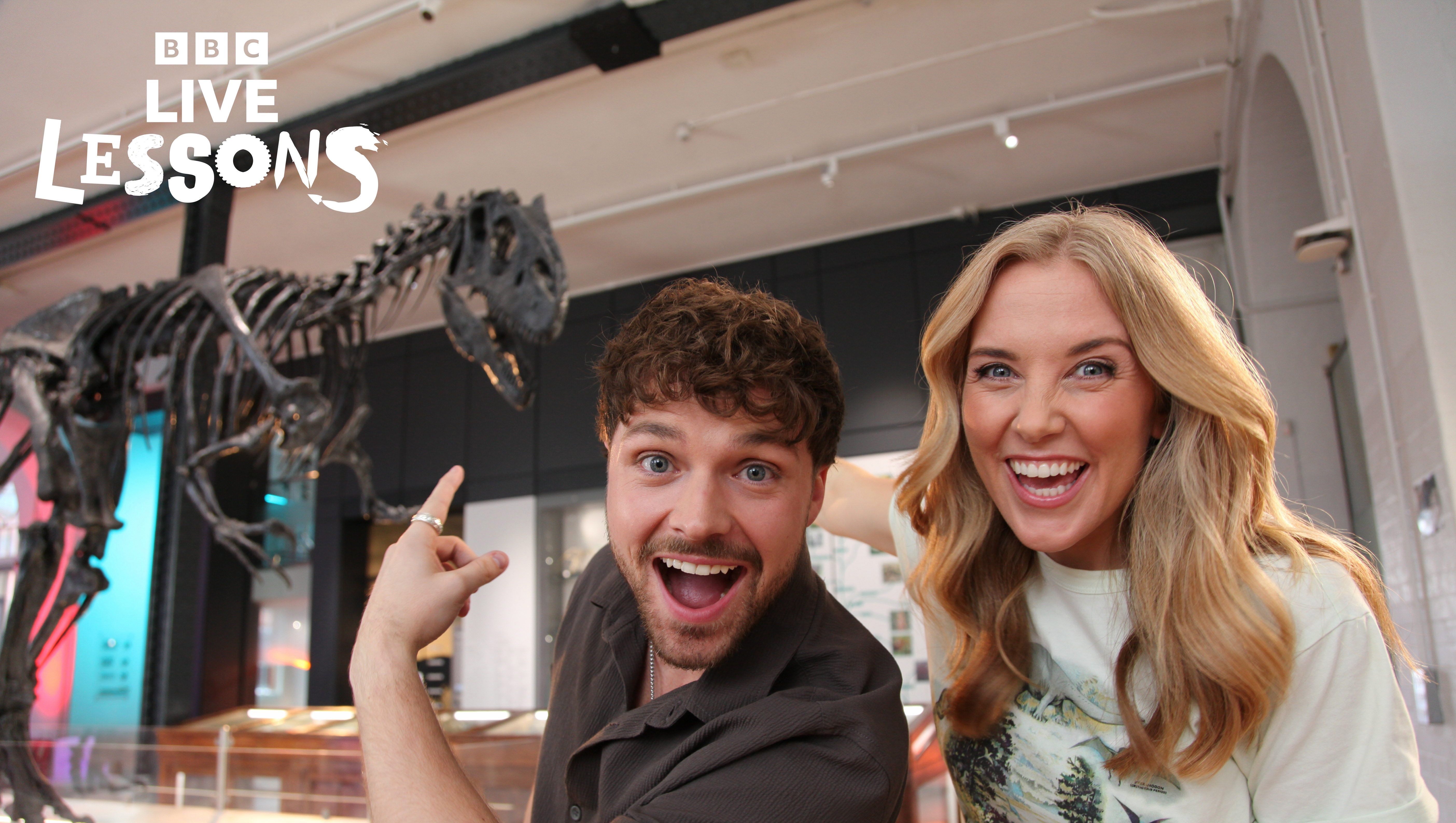 Presenters Maddie and Cam stand in amazement in front of an Allosaurus skeleton.
