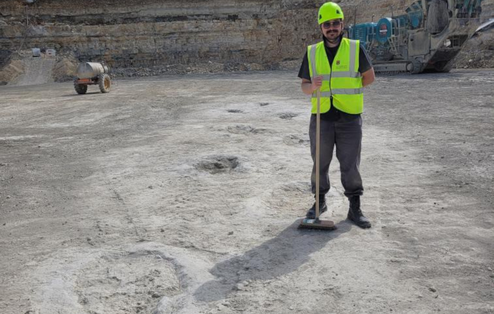 J. D. Dixon stands, broom in hand, alongside a fossil sauropod trackway.