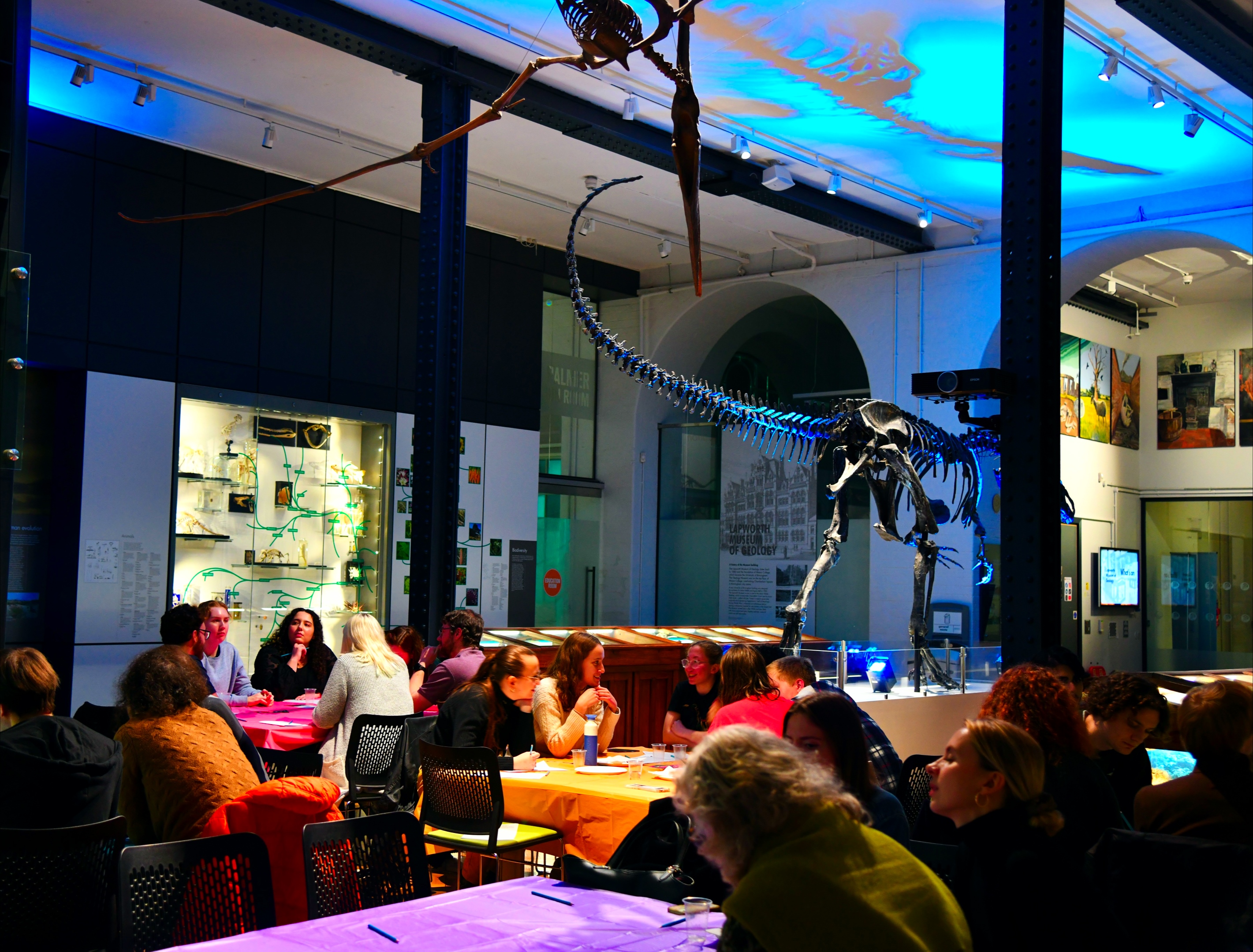 A crowd think over their answers to a quiz underneath a dinosaur skeleton and blue lights.
