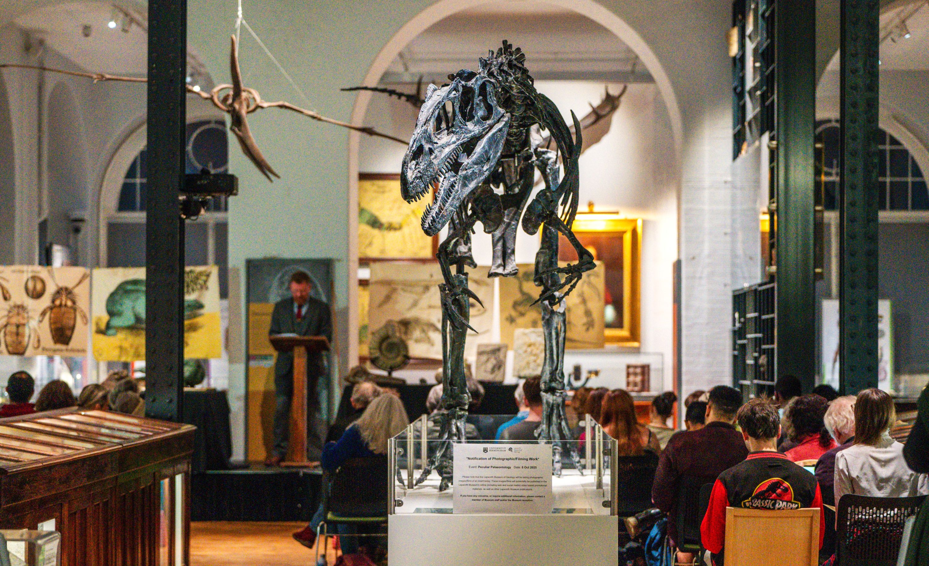 A group of people watch a geological lecture beneath a dinosaur skeleton.