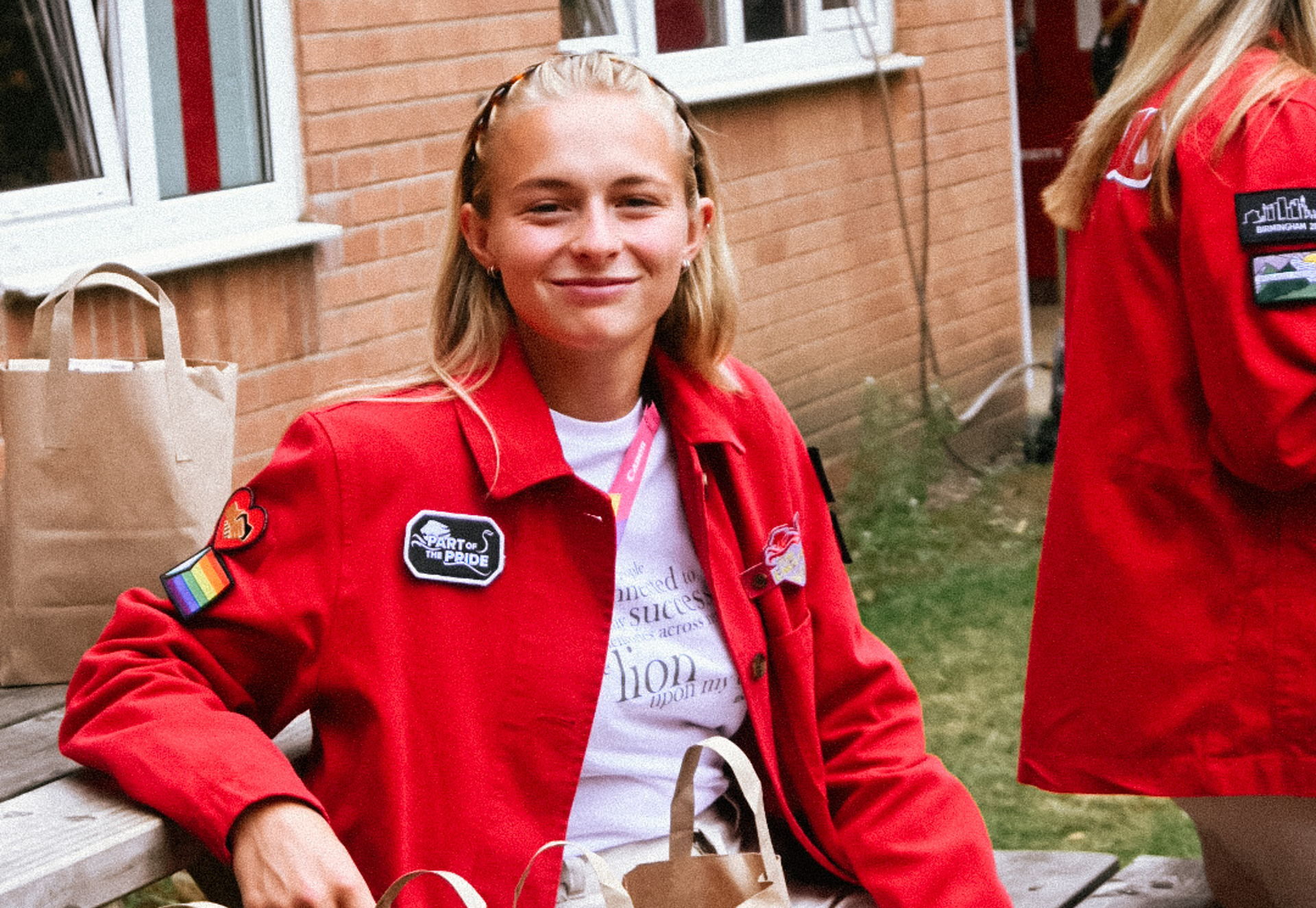 A young woman with blond hair and a red jacket sits on a bench, smiling at the camera.