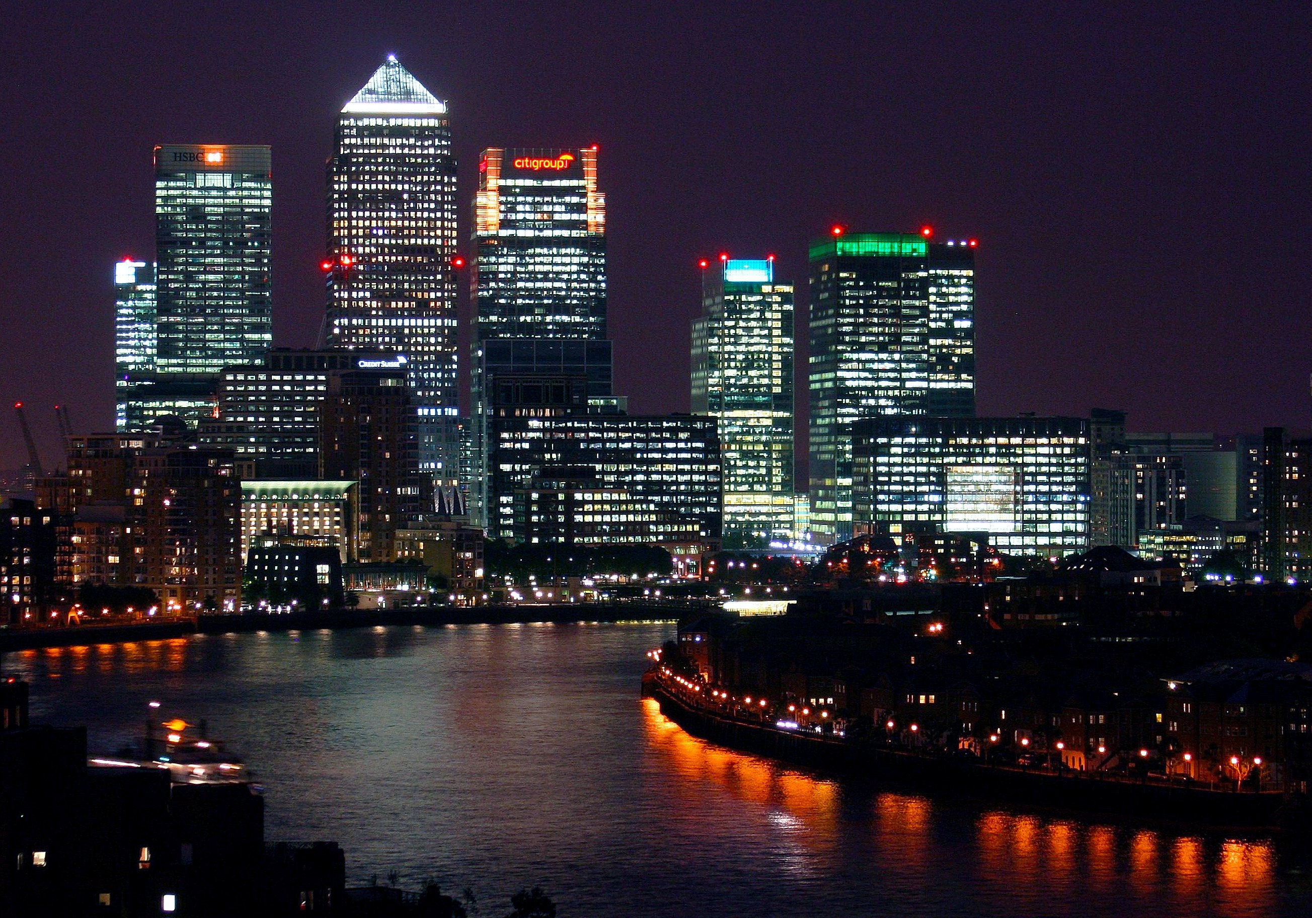 The city of London at night, with multicoloured lights shining from the skyscrapers of the financial district and reflected in the river below.