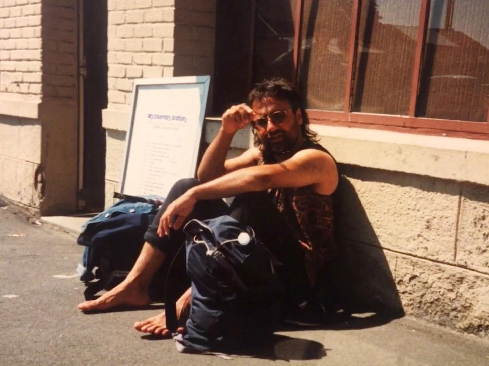 A young man with long hair and a beard, wearing glasses, sits barefoot in the sunshine accompanied by rucksacks