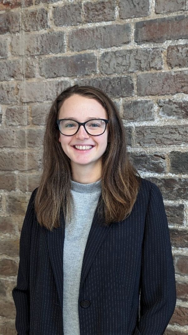 Morgan Roberts Profile Picture, stood by wall smiling, long hair and wearing glasses