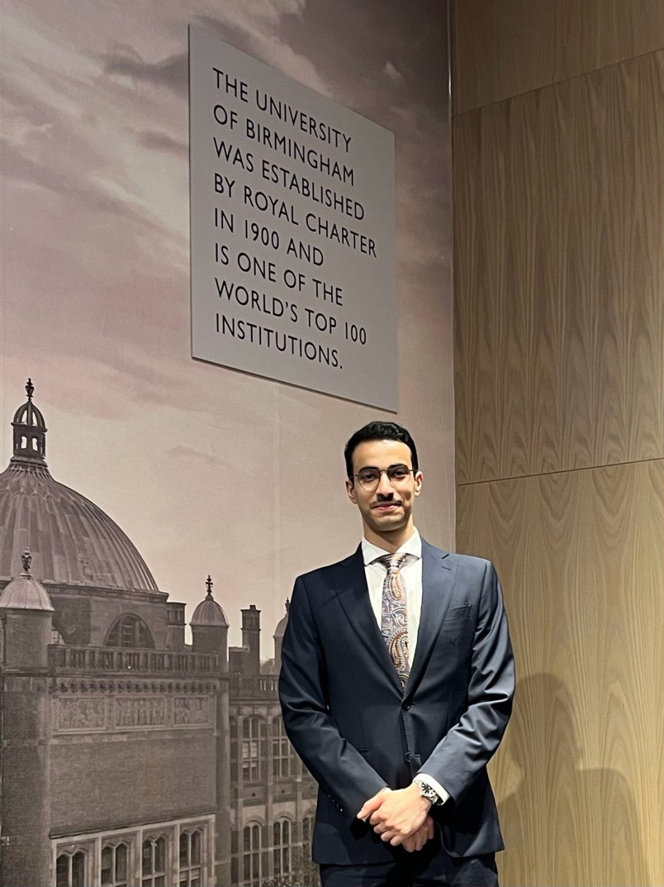 Muthana in a navy suit stood in front of a University of Birmingham image greyed out, inside the Aston Webb building