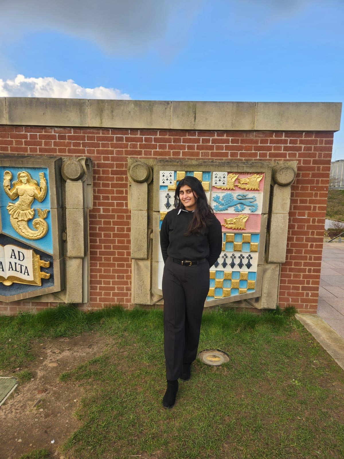 Nameera wearing black clothing smiling, stood in front of the Birmingham Crests on campus