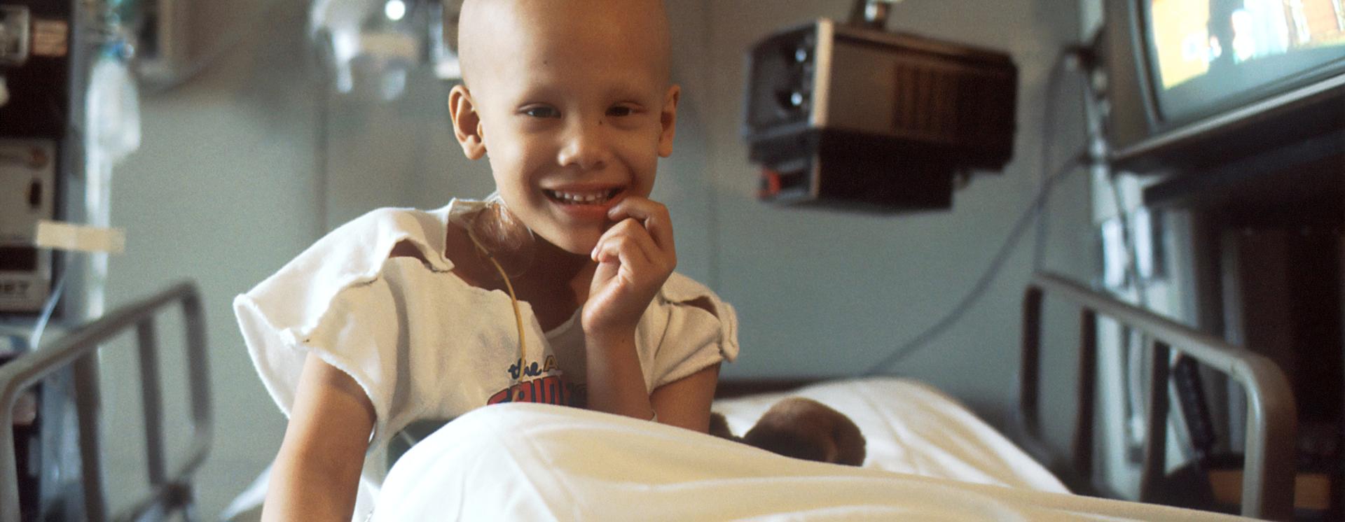 A child sat in a hospital bed smiling at the camera wearing a hospital gown and with medical equipment around them