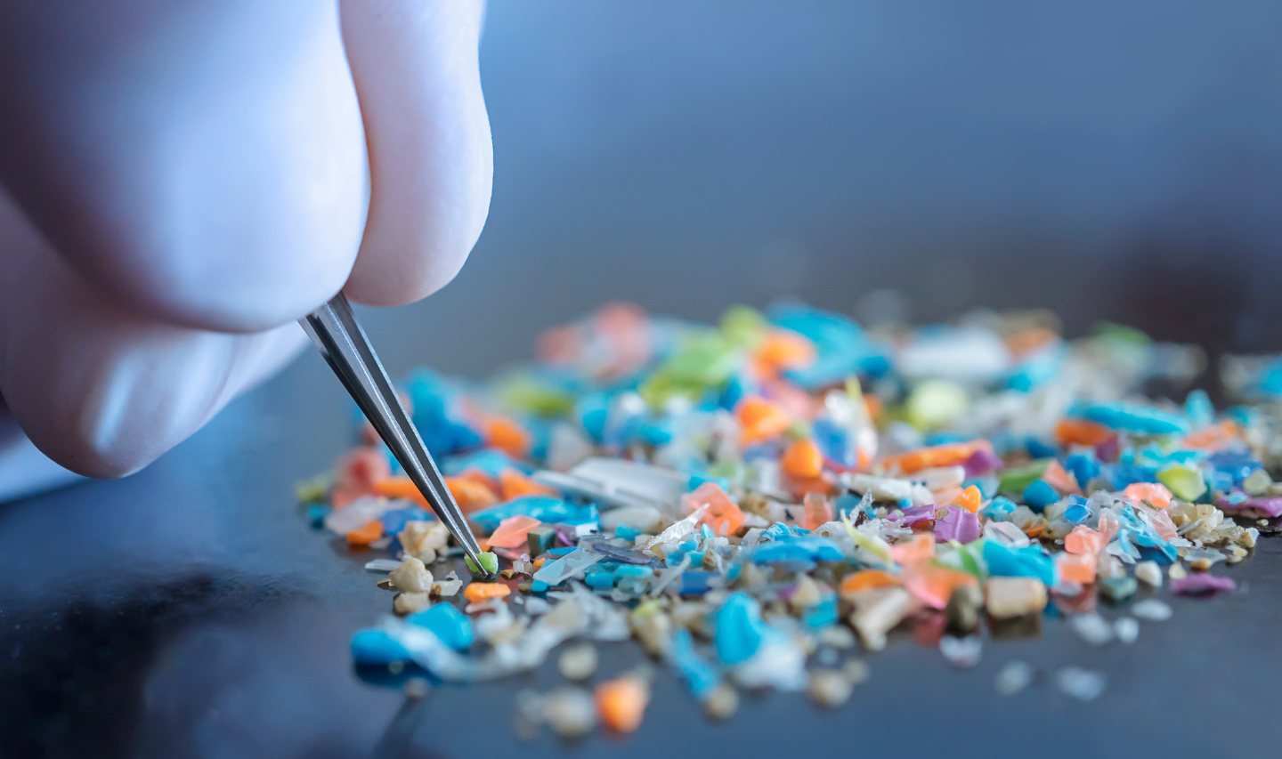 A gloved hand uses tweezers to pick up multi-coloured, microplastic particles from a table.