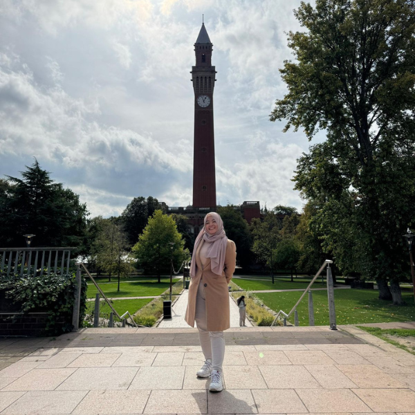 Noviana poses in front of Old Joe. She's wearing a light pink headscarf, a tan coat and white trousers.