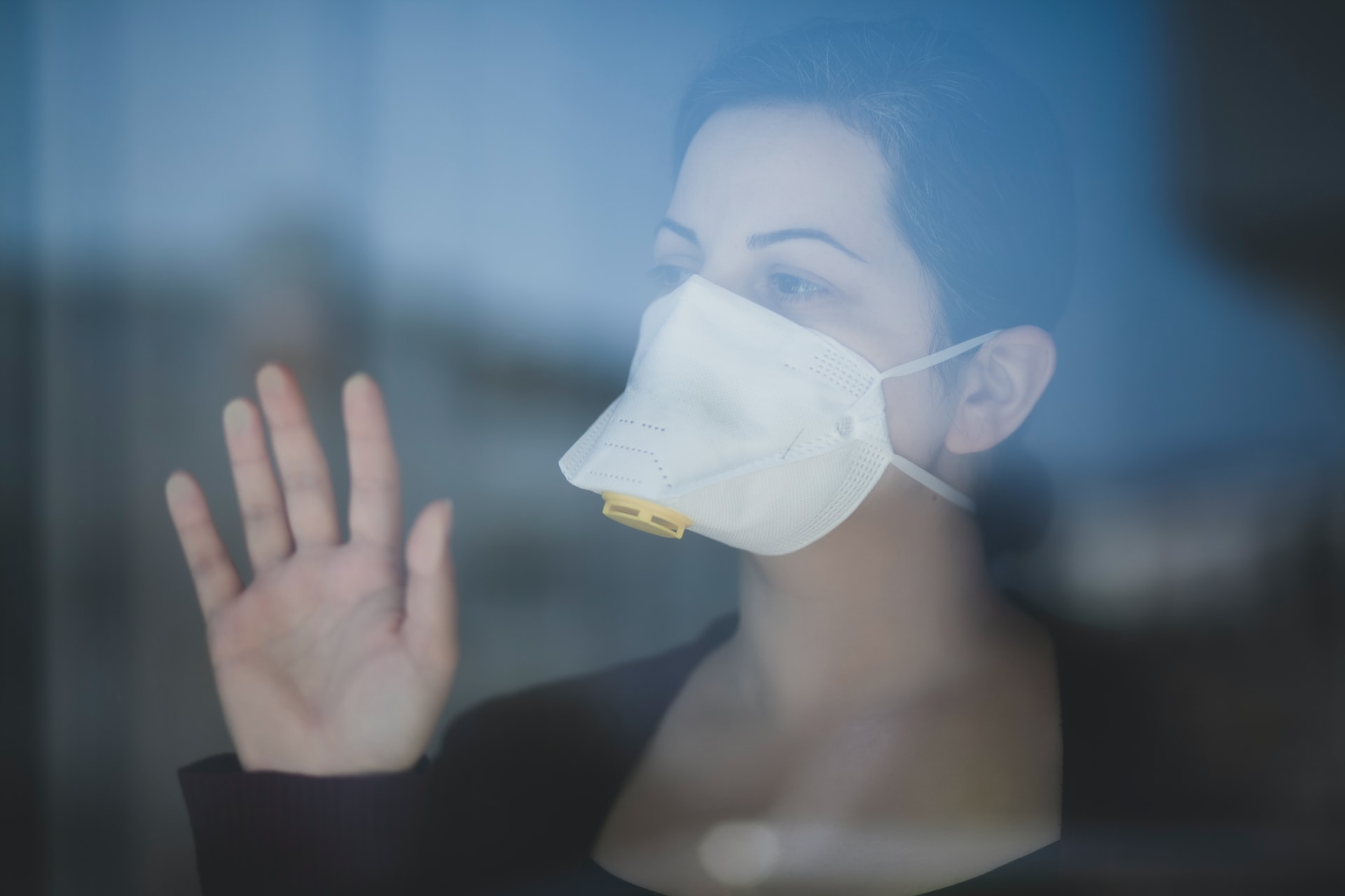 Woman wearing protective filter face mask standing behind pane of glass looking outside