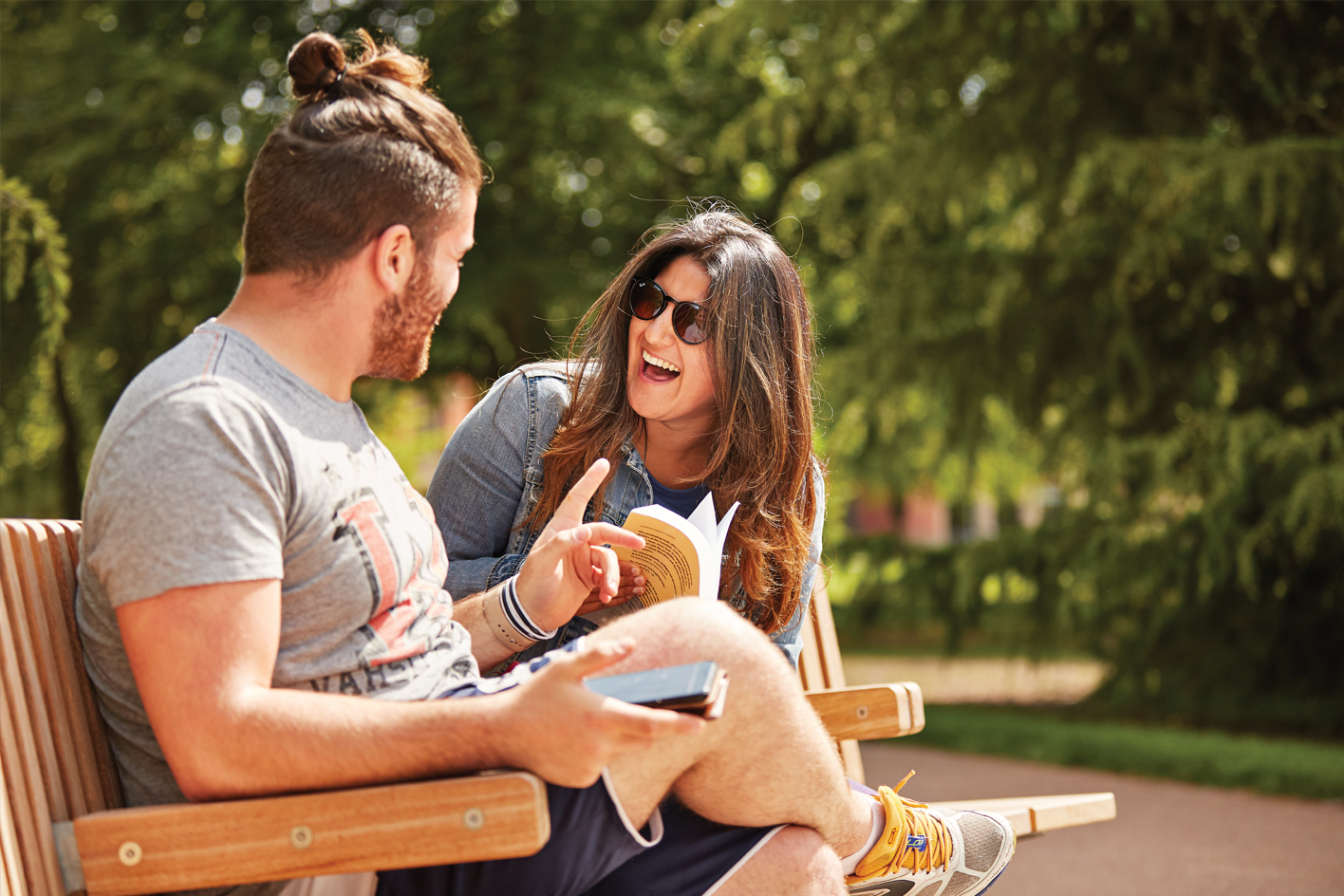 A male and female student sitting on a bench in the sunshine laughing together
