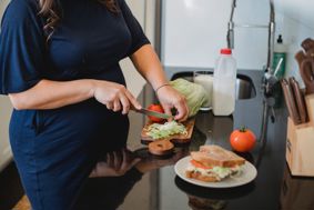 Pregnancy woman wearing blue dress chopping salad on wooden chopping board as part of food preparation