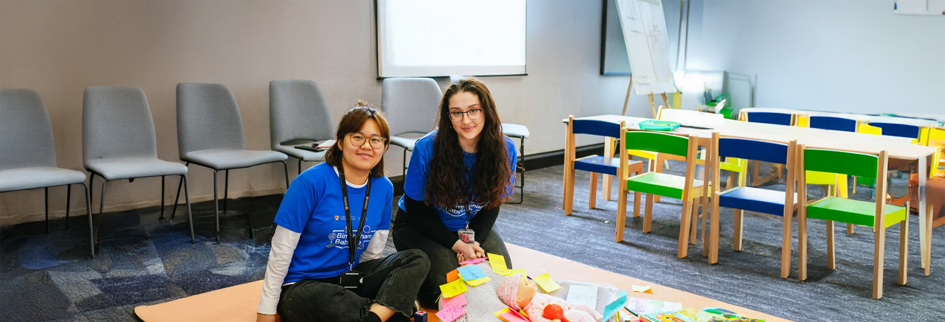 Two young women in a classroom setting sitting behind a crib with a baby doll in it