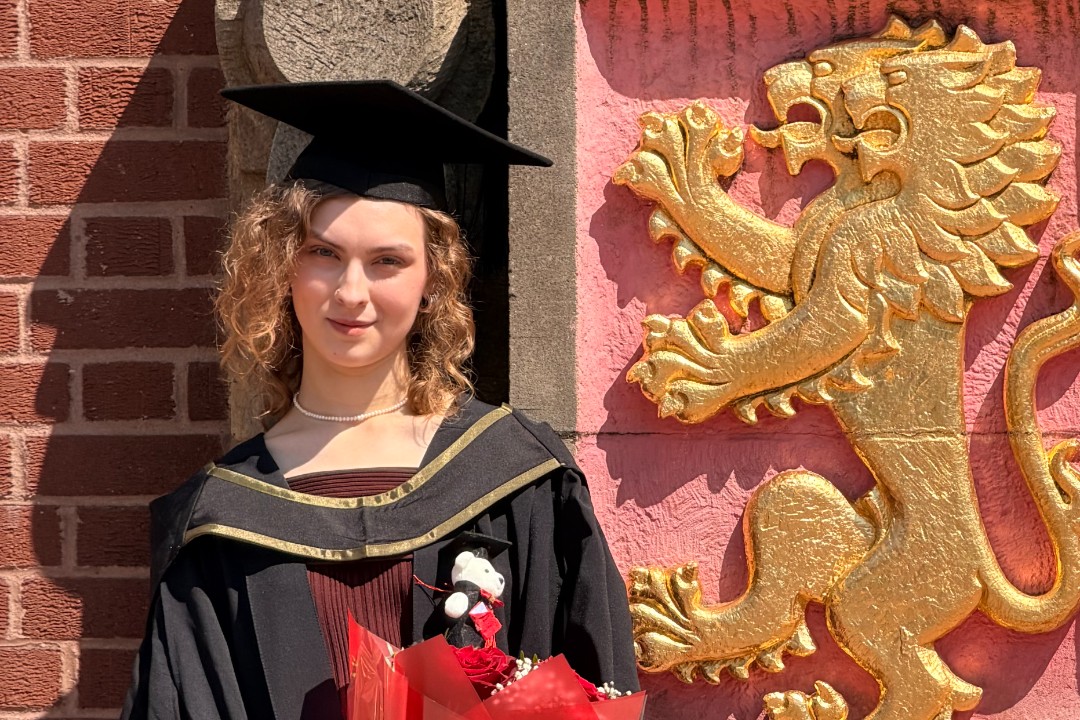 A woman in graduation cap and gown stands in front of the University of Birmingham Crest