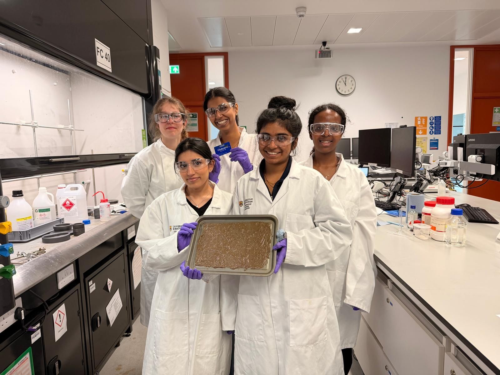 A group of students in white lab coats and protective glasses are stood in a laboratory with their project