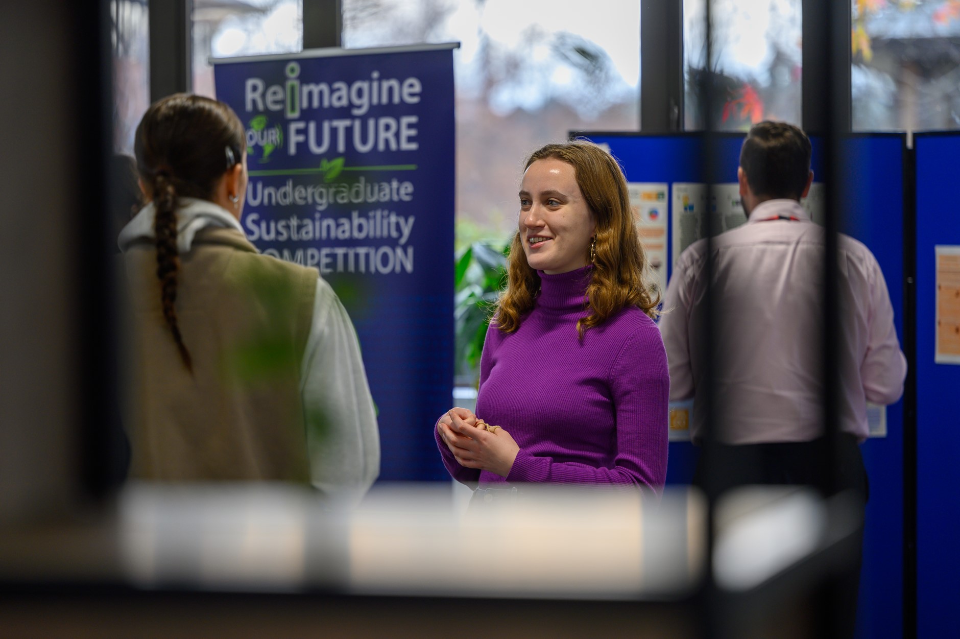 A woman in a purple top smiles while talking to another person, with a sustainability competition poster behind her.