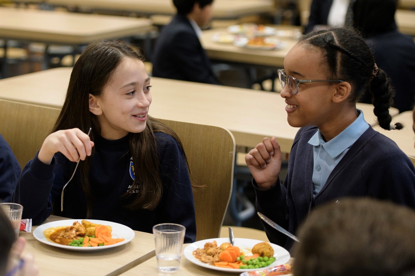 Two schoolgirls sit next to each other at a lunch table at school, talking and laughing with each other. In front of them is a healthy lunch with vegetables.