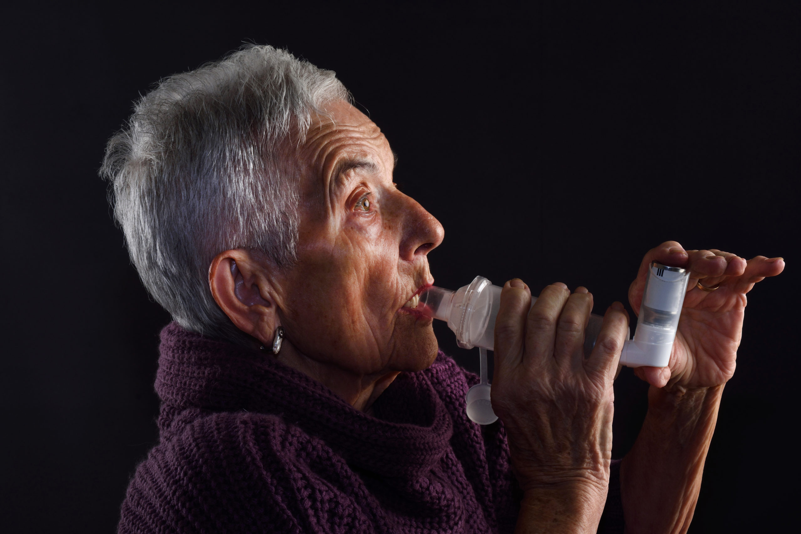 A woman with short grey hair and purple jumper using an asthma inhaler and spacer