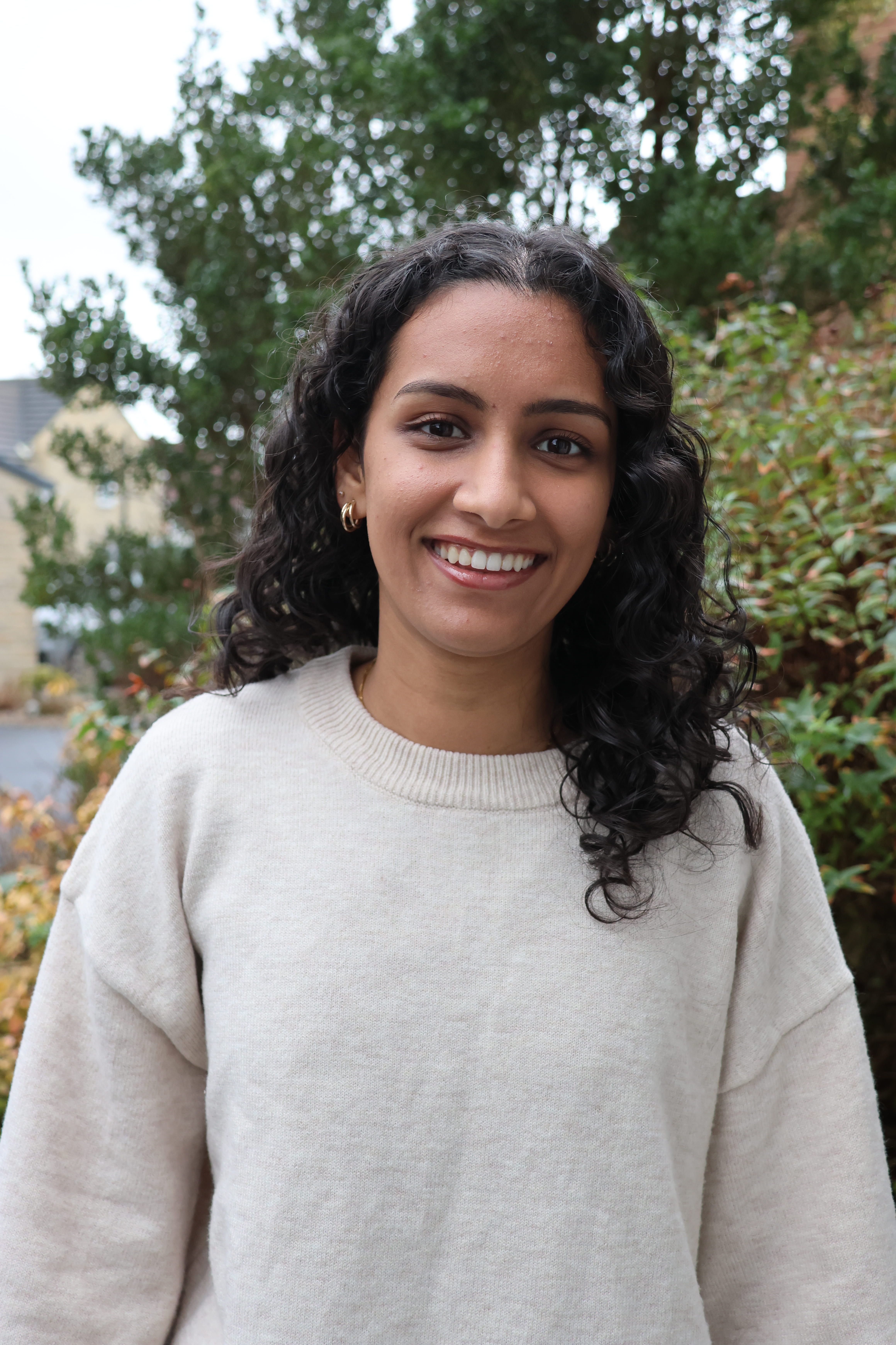 Shreya poses in front of a tree, smiling. She has curly black hair and is wearing a cream jumper.