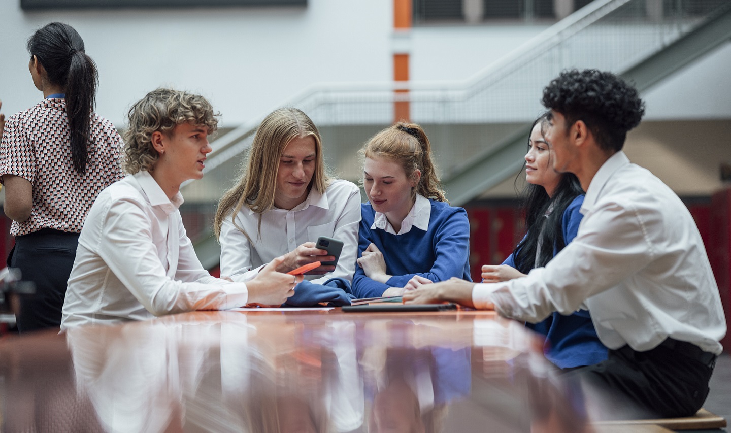 Five secondary school students sat round a table in school with two looking at a mobile phone