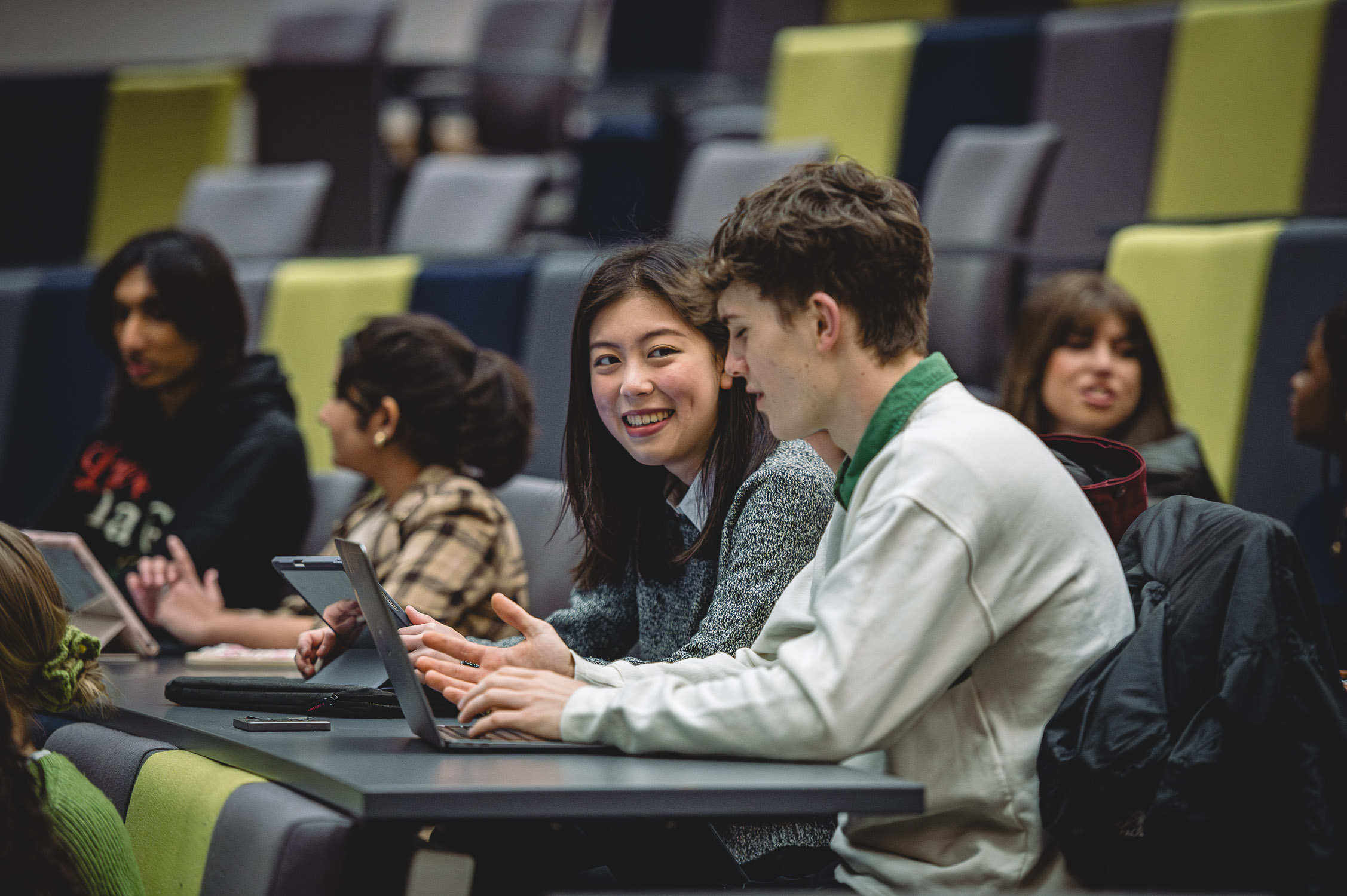 Two students chat in a lecture theatre, surrounded by other students