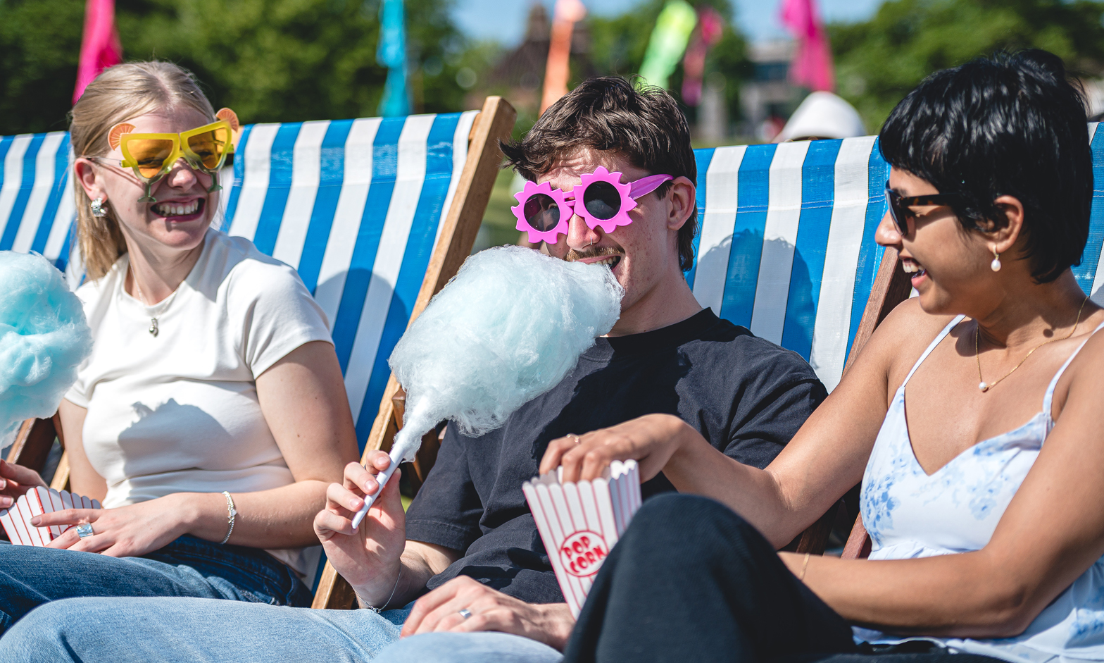 Three students sitting in deck chairs eating blue candy floss and popcorn while wearing novelty glasses