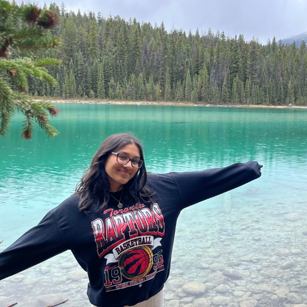 Tanisi is posing in front of a clear lake surrounded by a pine forest. She's wearing a Toronto Raptors jumper.