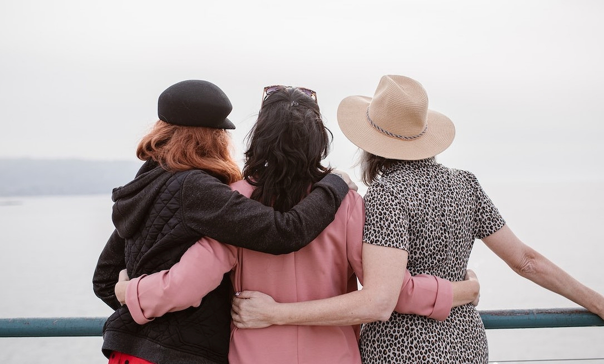 Three women with their backs to the viewer, look out to sea. They have their arms around each other.