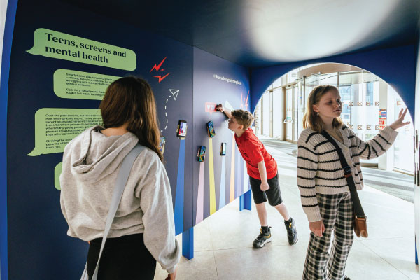 Three children interact with an exhibit in a blue tunnel under the title "Teens,s creens and mental health."