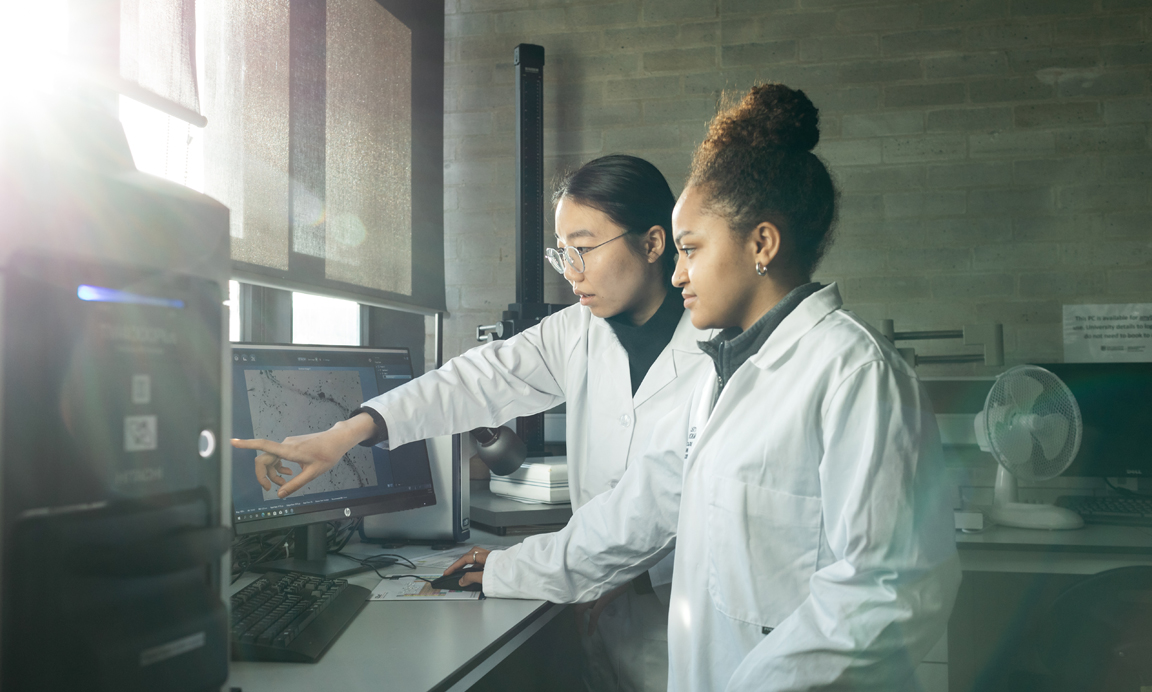 Two female students in lab wearing white coats pointing at a monitor