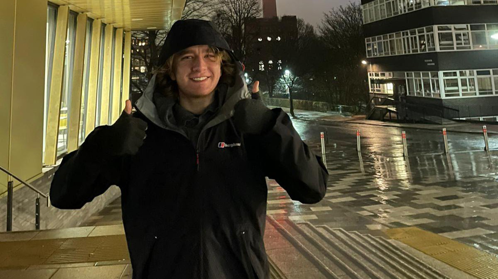 A student in a cagoule holds two thumbs up while getting rained on