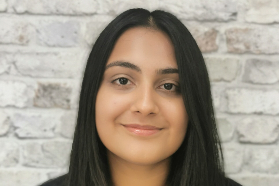 Jess, a female student with long black hair, smiles at the camera in front of a white brick wall