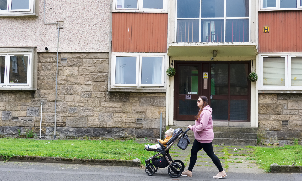 Young mum walks past flats pushing a baby in a buggy
