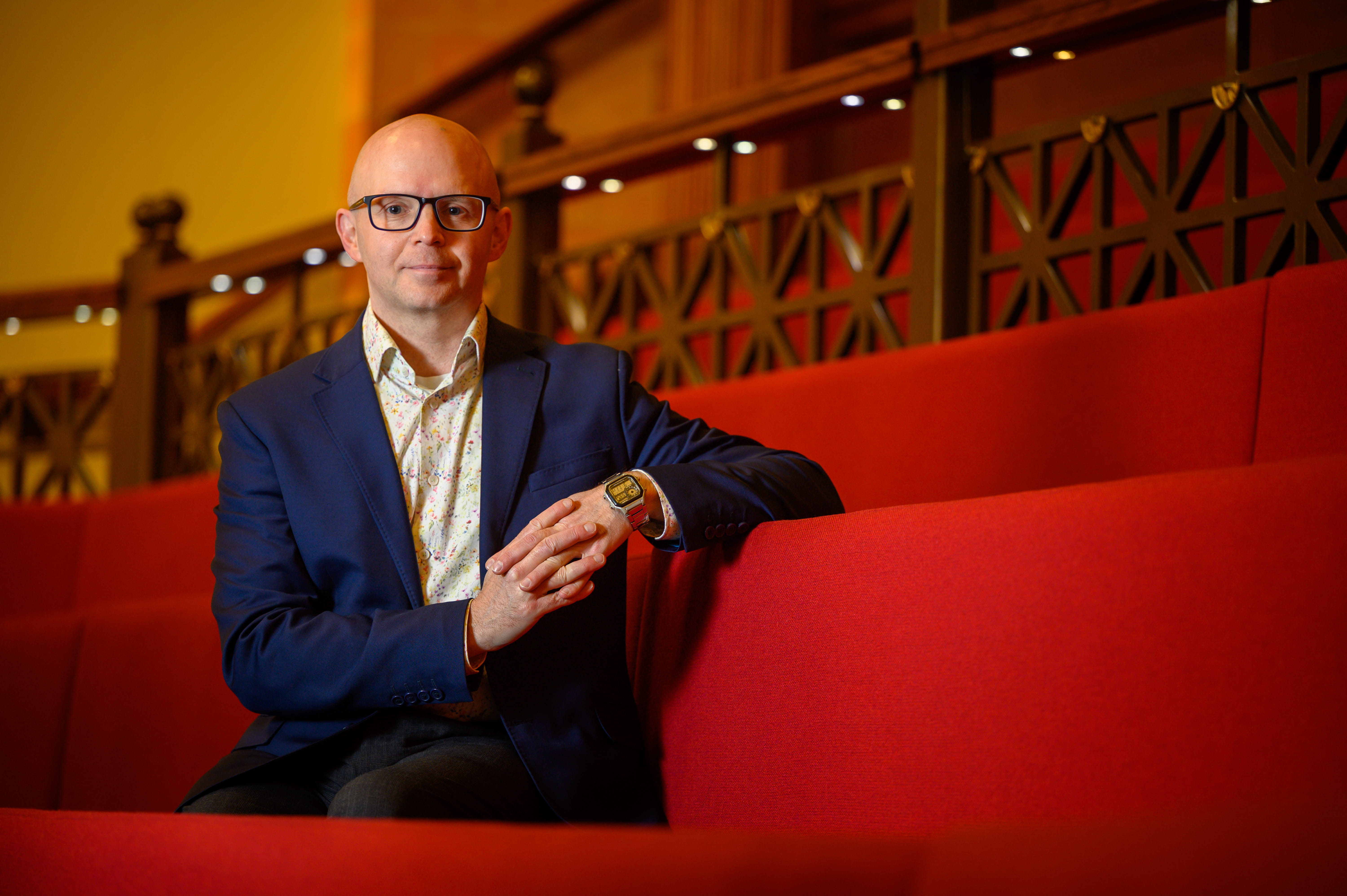 A man wearing glasses and a navy blazer sits on a red bench, resting his hands together.