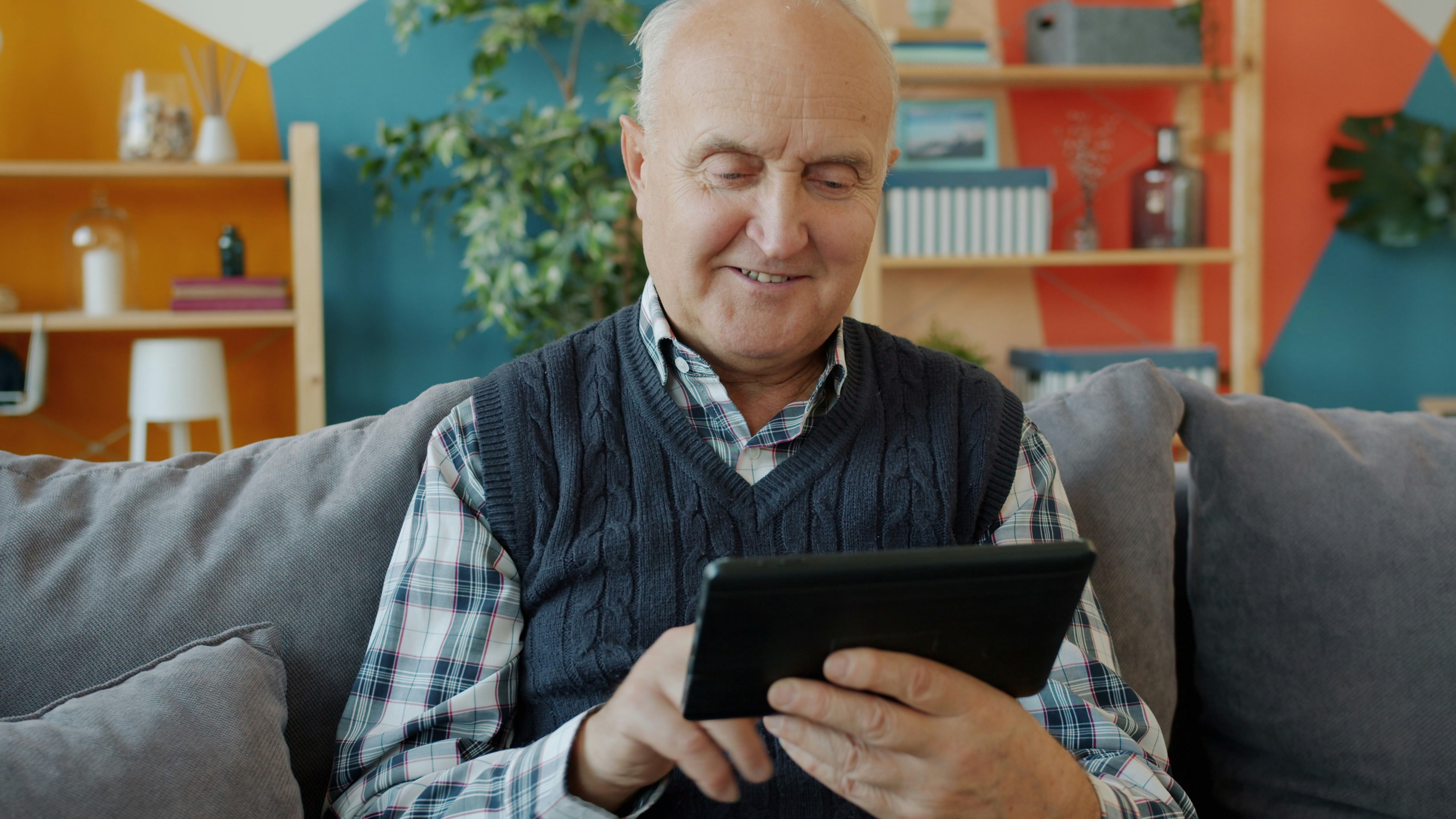 Cheerful senior man is swiping tablet screen enjoying online content sitting on couch at home. 