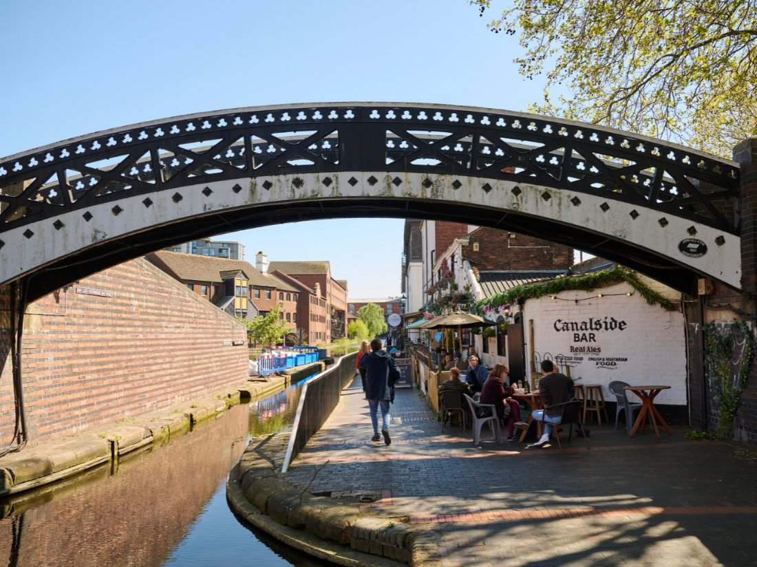 Black and white bridge crosses a canal and path alongside, with canalside bars and seating