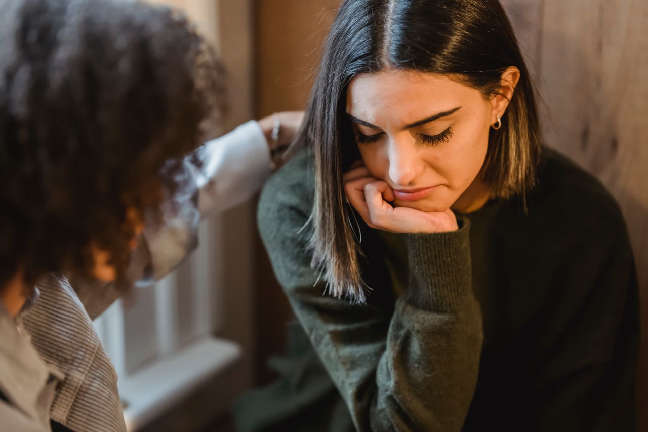One woman looking sad, with a friend offering support and putting an arm around her.