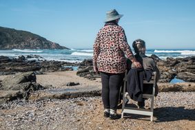 Two older women on the beach looking at the sea. One sat on a chair and one standing.