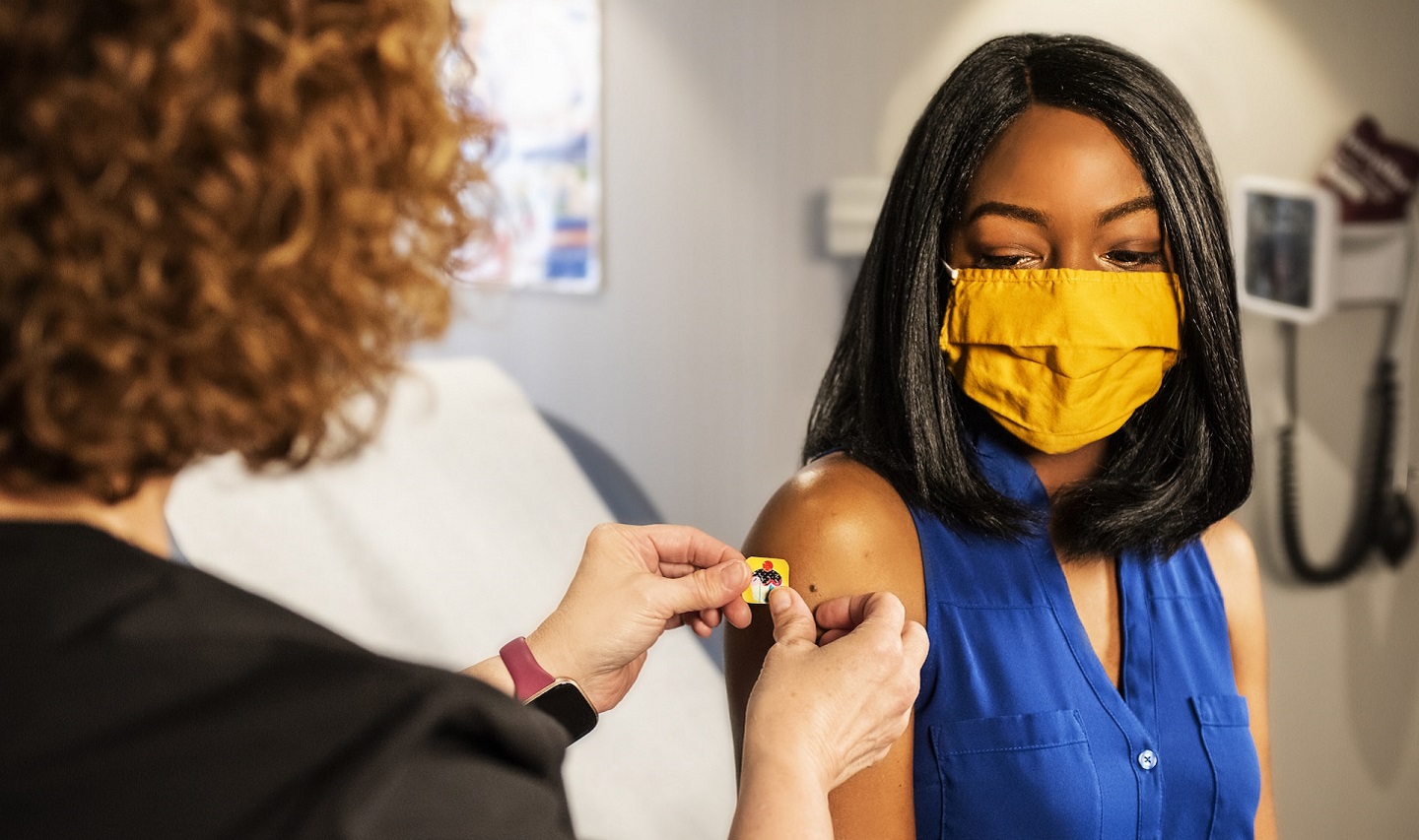 Young woman wearing facemask having sticker applied to arm after vaccination