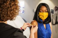 Young black woman wearing yellow face covering having plaster applied to vaccination site on upper right arm by doctor