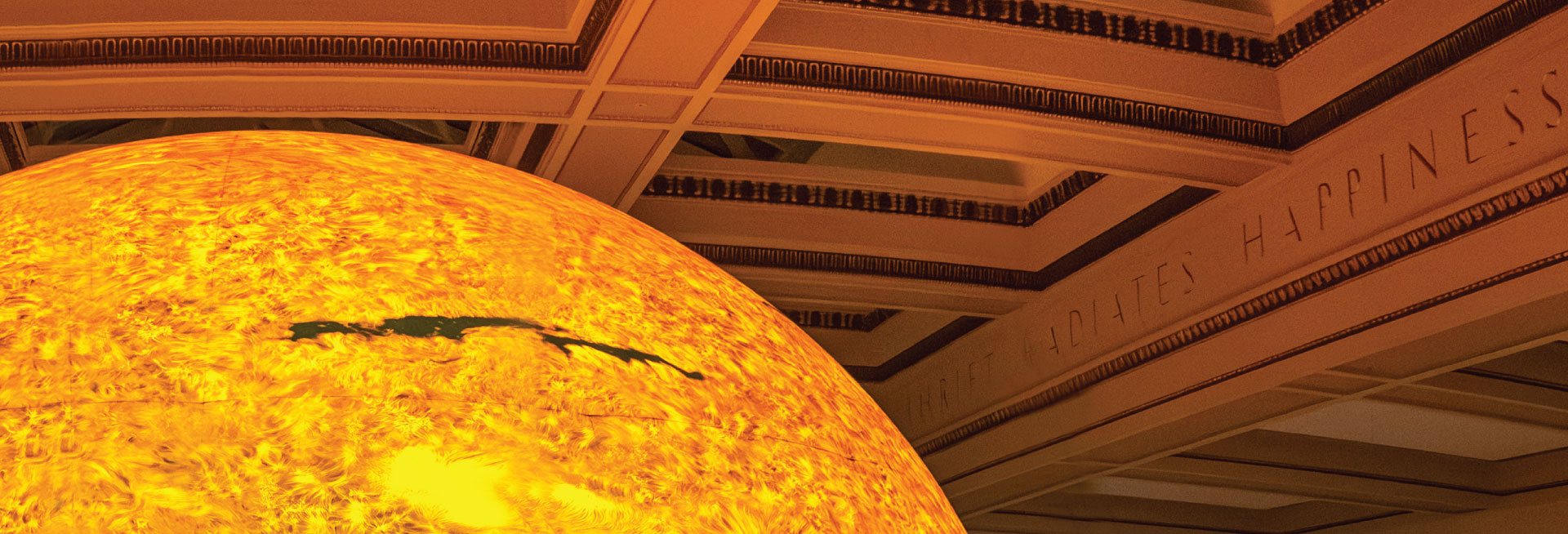 A portion of a large sculpture of the sun hanging from a ceiling engraved with the words "Thrift Radiates Happiness".