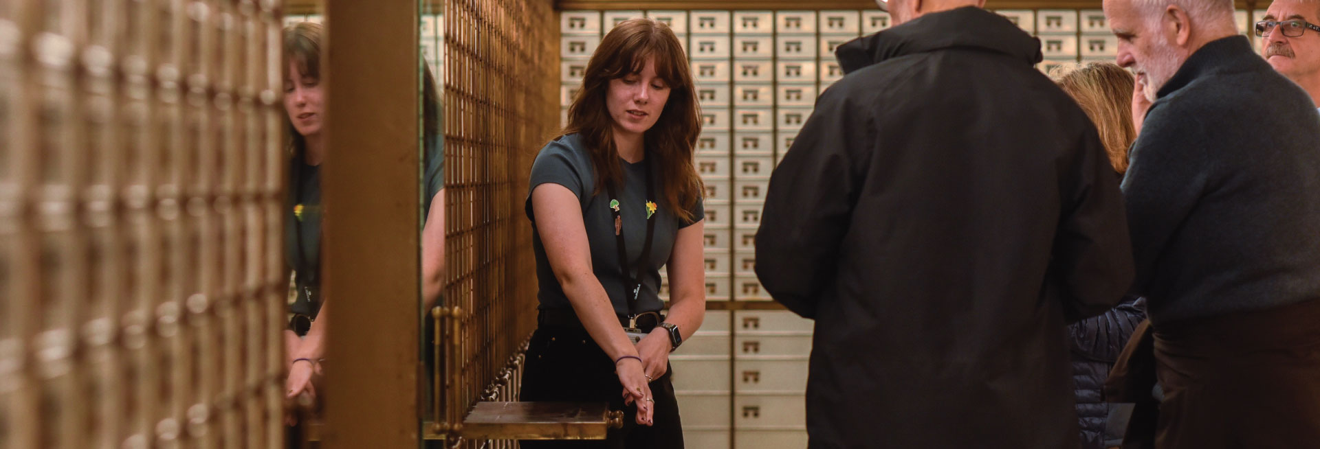A female guide gestures to a wall of safety deposit boxes in front of a tour group.